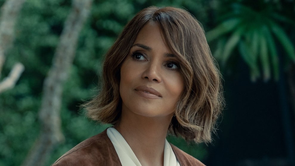 Halle Berry in a still from "Crime 101," looking off to the side with a thoughtful expression, wearing a brown suede jacket over a cream top, set against a blurred green jungle backdrop.