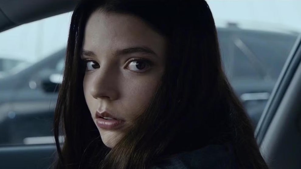 A young woman sitting in a car passenger seat looks over her shoulder and directly into camera with a concerned expression