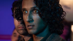 A vampire character with glowing amber eyes and dark curly hair gazes intensely forward under dramatic teal lighting, with a second figure blurred in the background.
