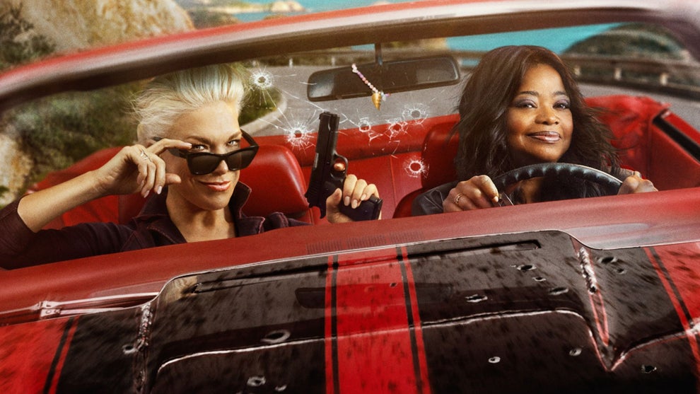 Two women ride in a red convertible with a bullet-riddled windshield — one in the passenger seat tips her sunglasses down while holding a handgun, while the other smiles confidently at the wheel, with a coastal landscape visible behind them.