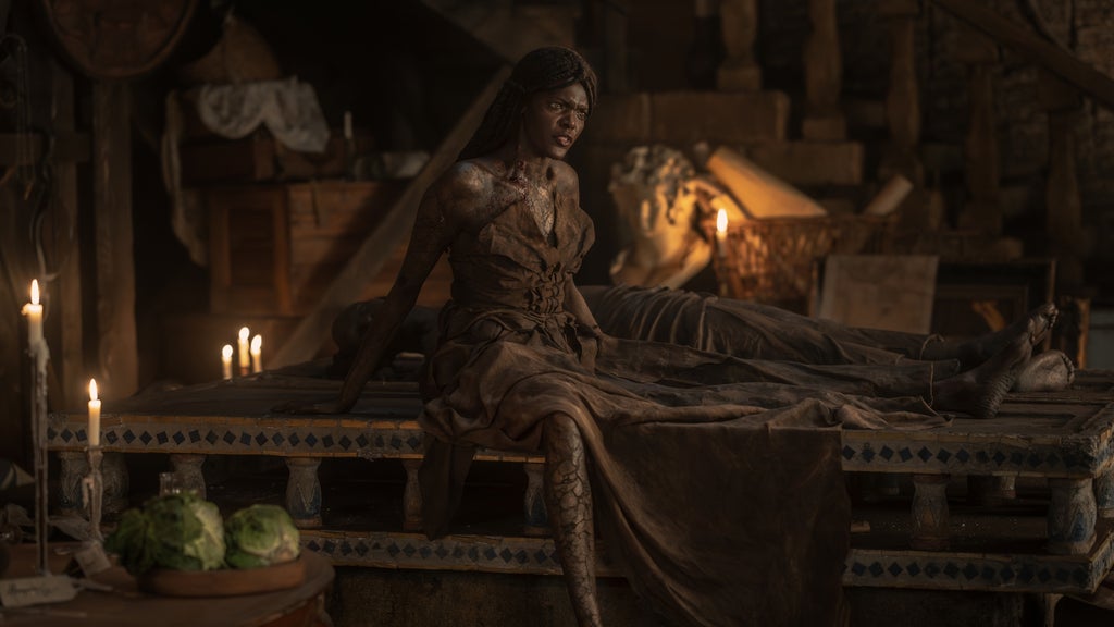 A vampire character with cracked skin and blood at her neck sits upright on an ancient stone bier surrounded by candlelight, with a prone figure lying beside her in a dimly lit, torch-lit chamber.