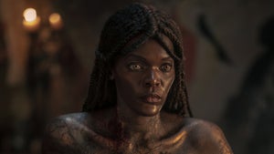 A close-up of a vampire character with glowing amber eyes, locs pulled back, and cracked golden-dark skin marked with blood at the collarbone, staring with cold intensity in candlelit darkness.
