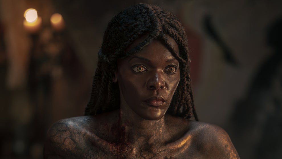 A close-up of a vampire character with glowing amber eyes, locs pulled back, and cracked golden-dark skin marked with blood at the collarbone, staring with cold intensity in candlelit darkness.