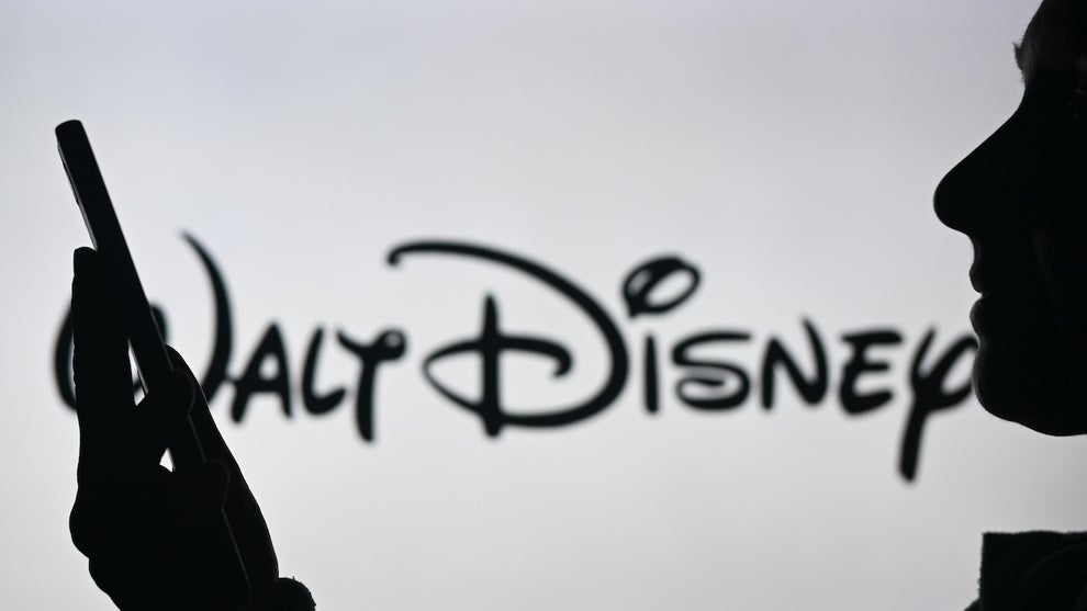 An image of a woman holding a cell phone in front of a Walt Disney logo displayed on a computer screen (Credit: by Artur Widak/NurPhoto)
