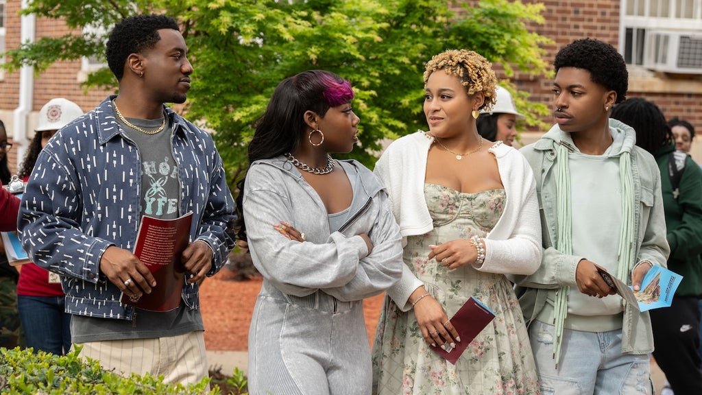 "Four young adults stand together outdoors on a college campus, dressed in casual and semi-formal clothes, holding pamphlets and talking, with trees, other people, and a brick building in the background."