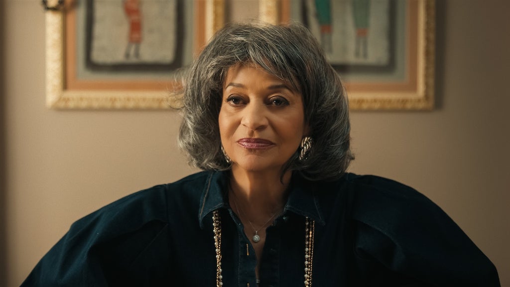 "Woman with gray hair wearing a dark blouse and gold necklace, sitting indoors in front of framed artwork on a beige wall, softly lit room."
