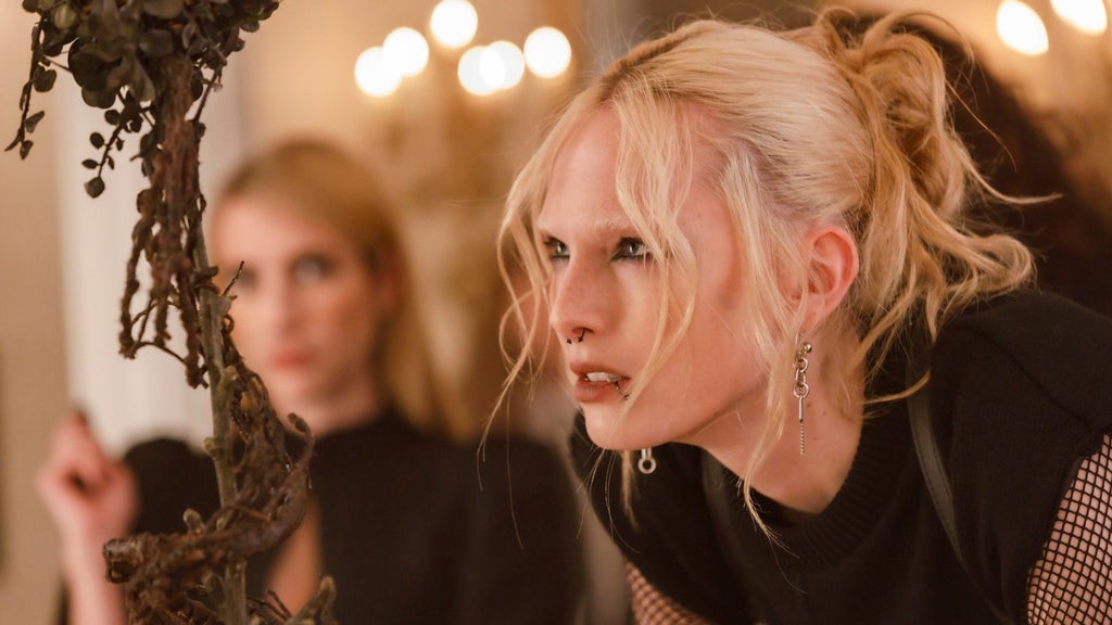 Blonde woman with a nose ring, multiple earrings, and lip piercing leans toward a dried branch, another blonde figure blurred behind her, candlelight in the background.