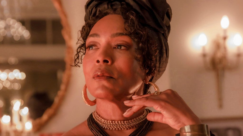 A woman in a dark head wrap, large gold hoop earrings, and layered beaded necklaces touches her chin, softly lit by candlelight.