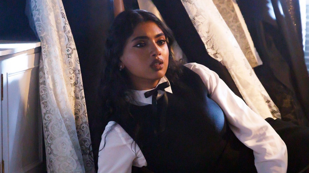 A woman with dark wavy hair, a black bow at her collar, and a white blouse under a black vest leans back among lace curtains, looking startled.