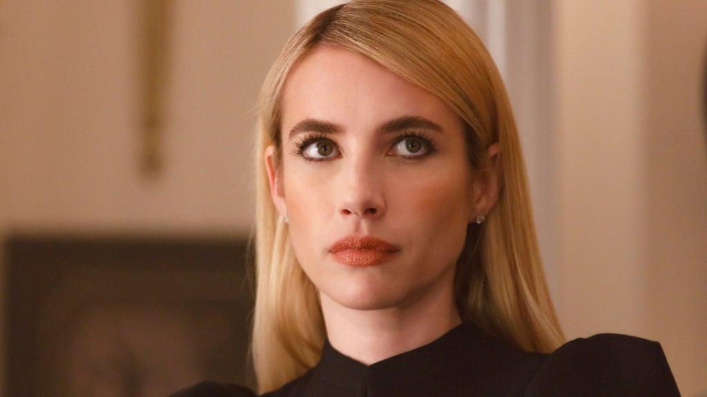 Close-up of a blonde woman with sleek straight hair, dark eyeliner, and orange lipstick, wearing a black high-collared top, looking upward with a serious expression.