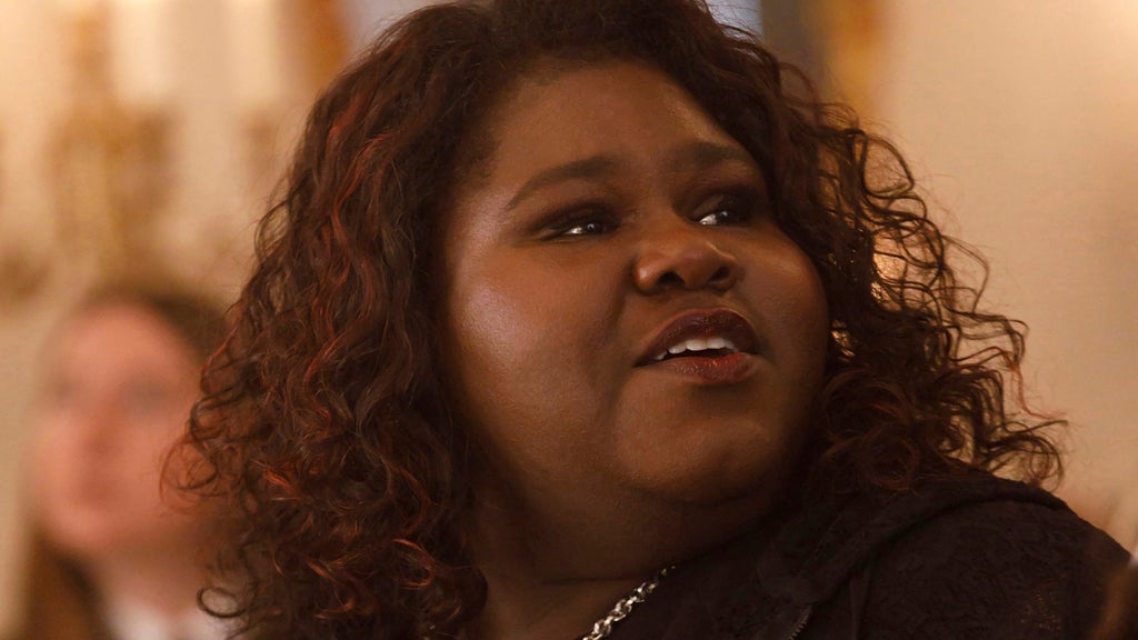 Close-up of a woman with curly dark hair and a chain necklace, looking up and to the side with an open-mouthed expression in a warmly lit room.