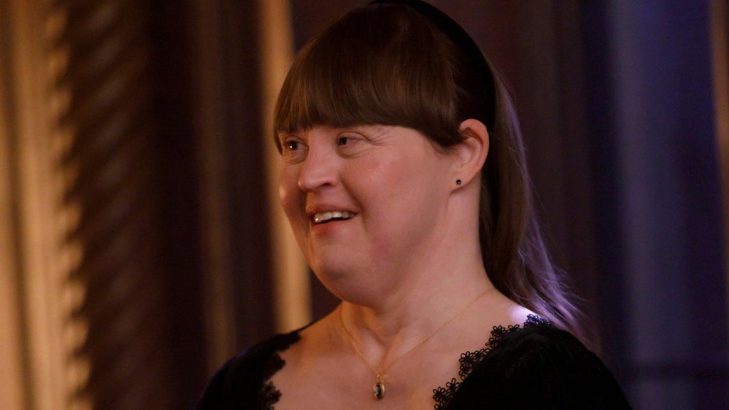 A woman with blunt-cut bangs and a headband smiles and looks to the side, wearing a black lace-trimmed top and pendant necklace.