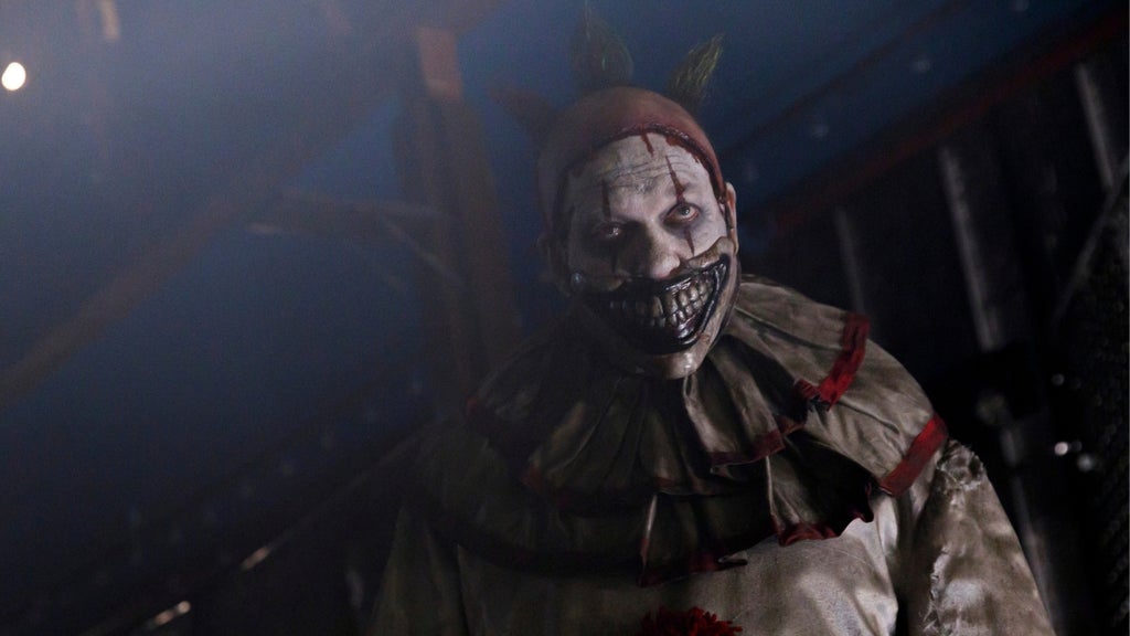 Menacing clown with a torn, stitched-looking mask-like grin, red headpiece, and tattered costume stands in dim, foggy blue light.