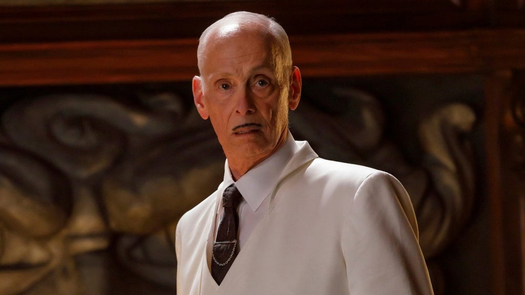 A bald man in a white suit and dark tie with a skull-shaped tie clip stands against an ornate carved wood panel, expression stern.