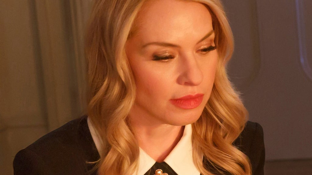 Close-up of a blonde woman with wavy hair and red lipstick, wearing a black blazer with white collar, looking downward.