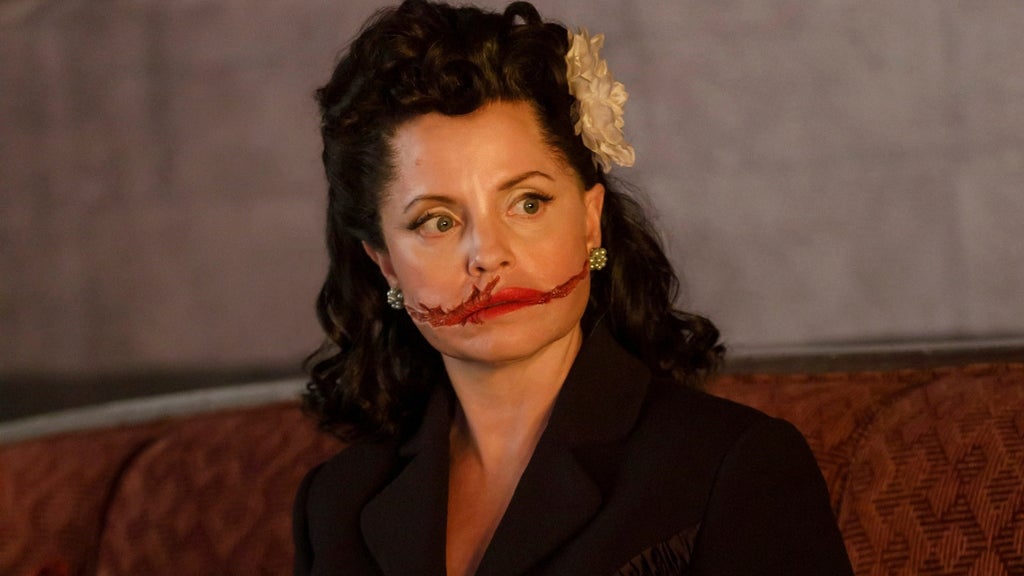 oman with dark curled 1940s-style hair and a flower hair clip has a deep red gash carved across both cheeks like a wide smile, wearing a black blazer.