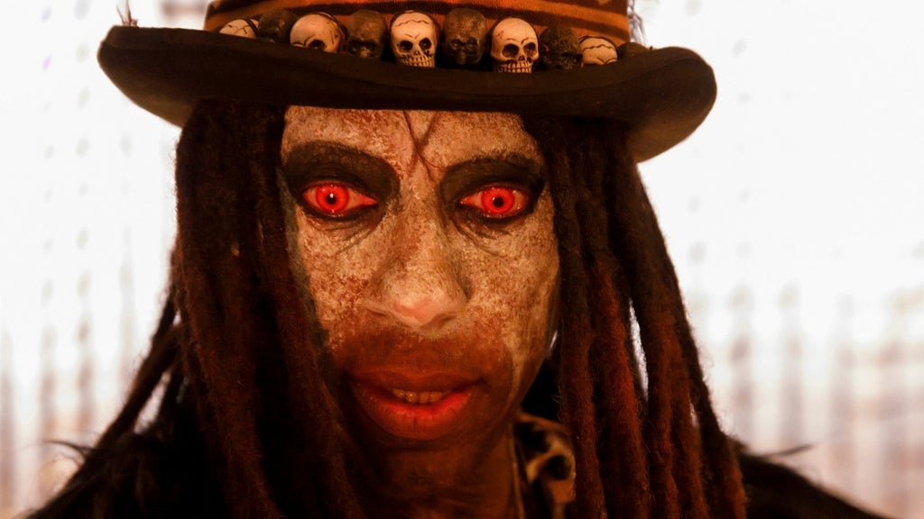 Close-up of a person with weathered face paint, red contact lenses, long dreadlocks, and a top hat adorned with small skulls, staring intensely at the camera with red eyes.