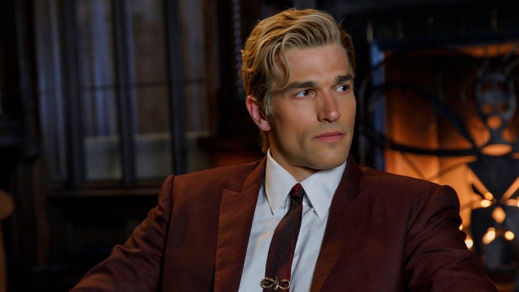 A mn with wavy blond hair in a maroon suit and dark tie sits in a firelit room, looking off to the side.