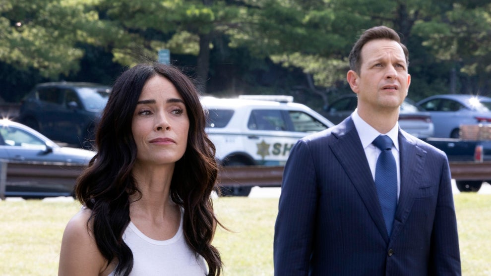 A woman with long, dark hair stands beside a man in a business suit stand outside in a sunny scene, both staring at something in the distance. Parked cars can be seen in the background behind them.