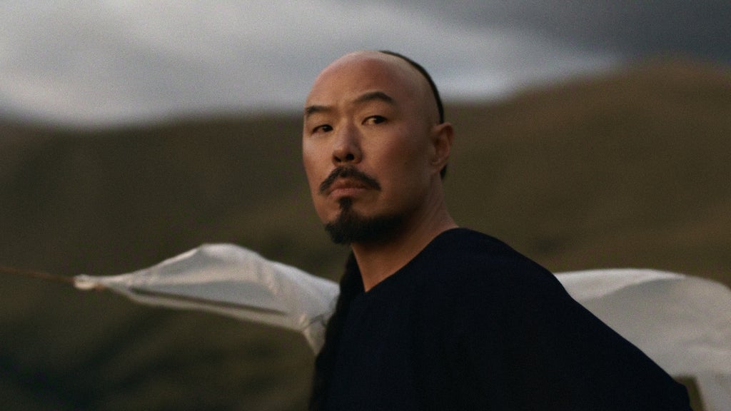 "A man with a mustache and goatee stands outdoors against rolling hills and cloudy sky, dressed in dark clothing with a white cloth behind him, looking toward the camera in a natural, rugged landscape."