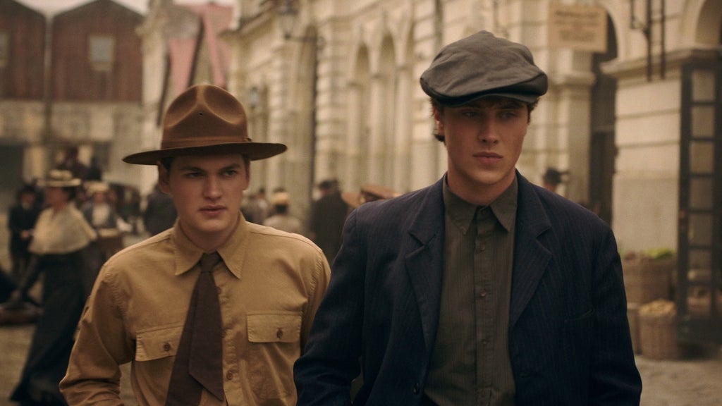"Two young men in vintage clothing and hats walk on an old city street with historical buildings and people in period attire in the background, evoking an early 20th-century urban setting."
