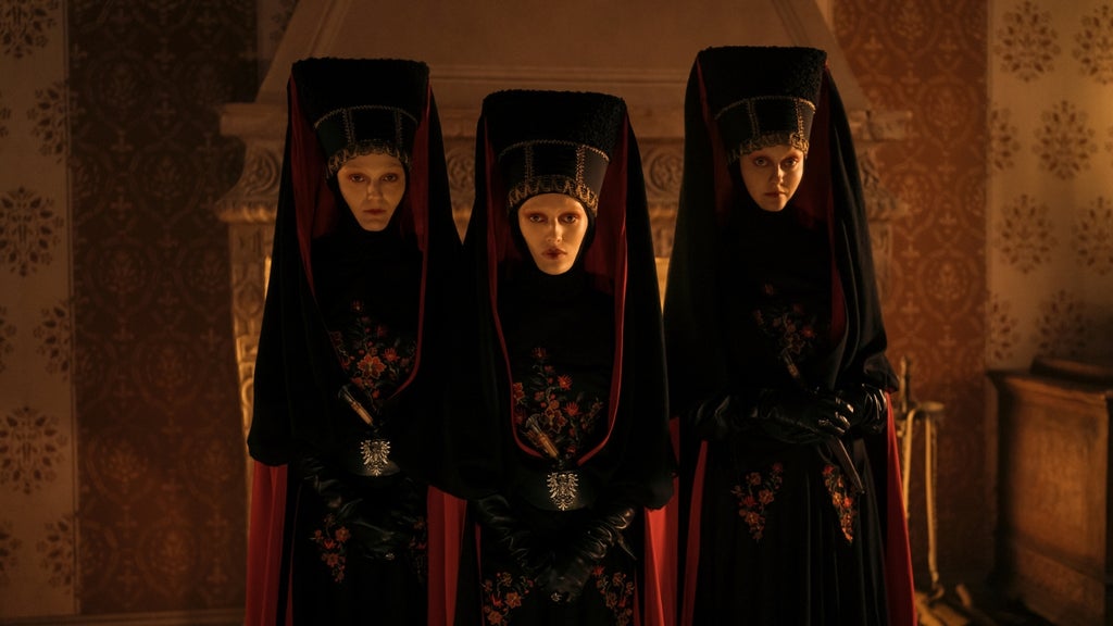 Three pale women in matching black fur headdresses and dark floral-embroidered robes with leather gloves stand side by side in a dim room.