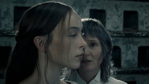 Two women stand close together in profile, one dark-haired in the foreground, against the backdrop of a crumbling stone building.