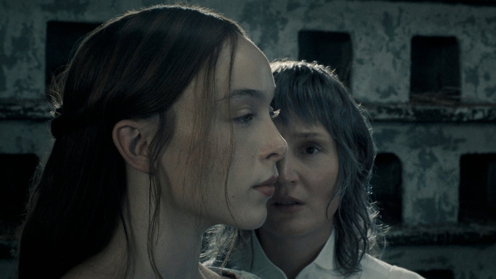 Two women stand close together in profile, one dark-haired in the foreground, against the backdrop of a crumbling stone building.