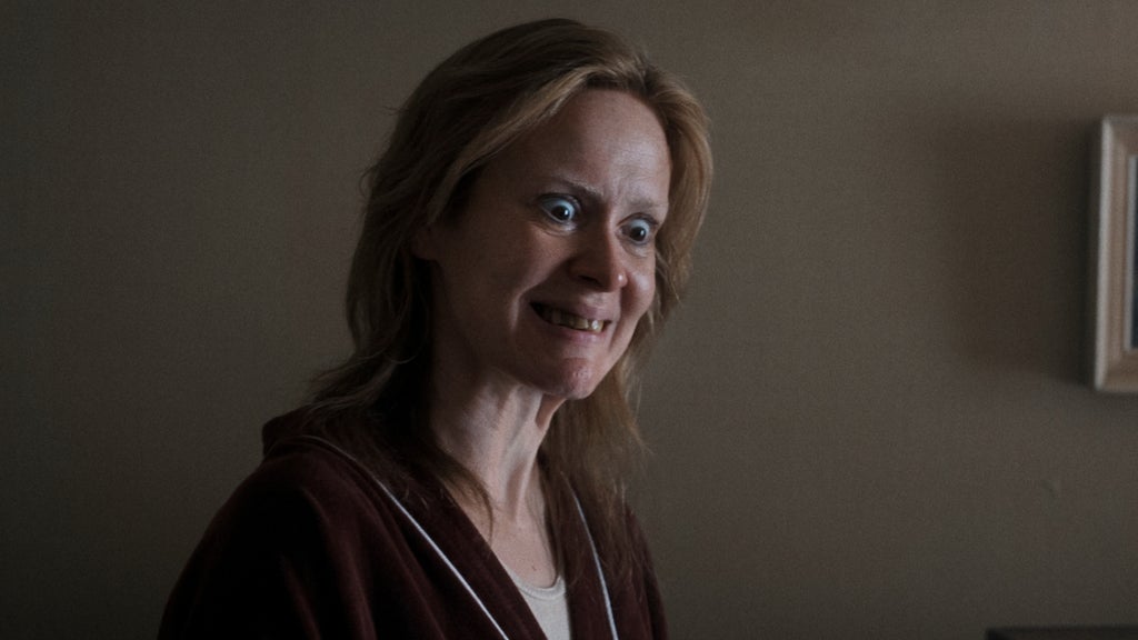 A woman with disheveled hair and wide, unsettling eyes smiles oddly in a dim room.