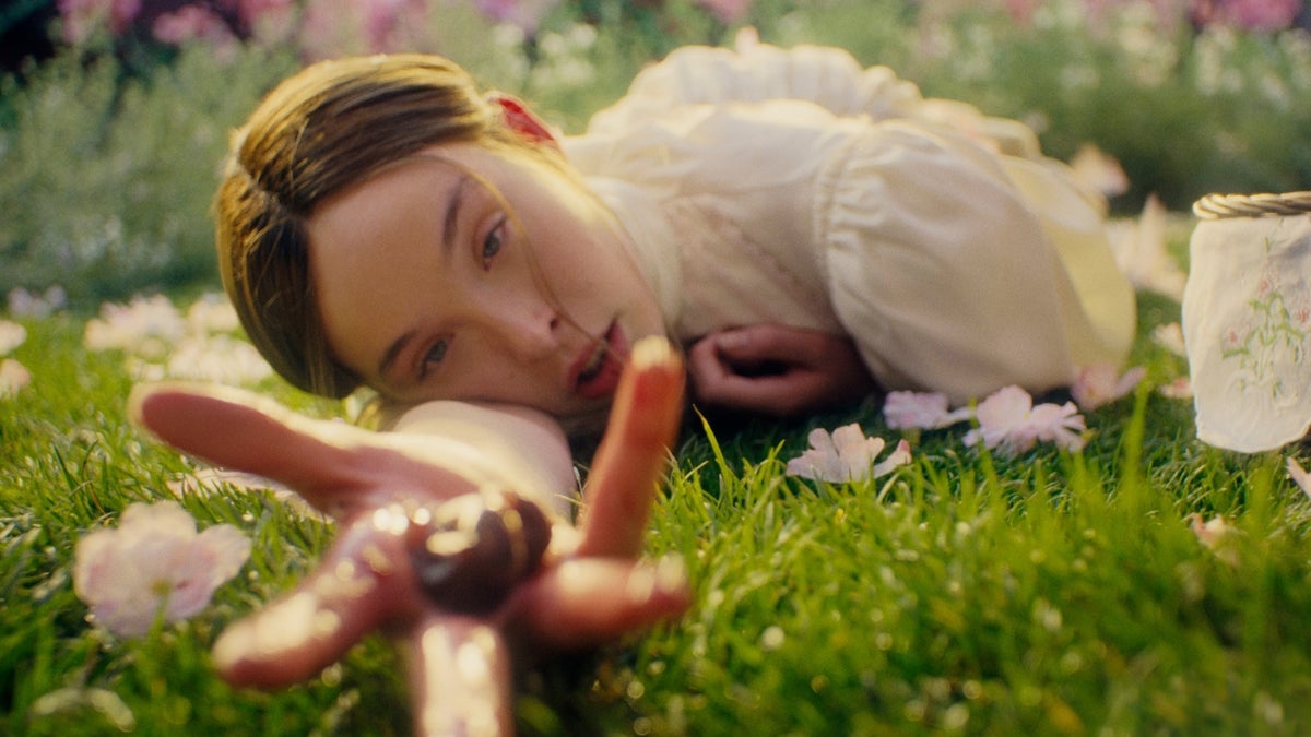 A young woman in a cream dress lies on grass among scattered flower petals, reaching toward the camera with something in her hand.