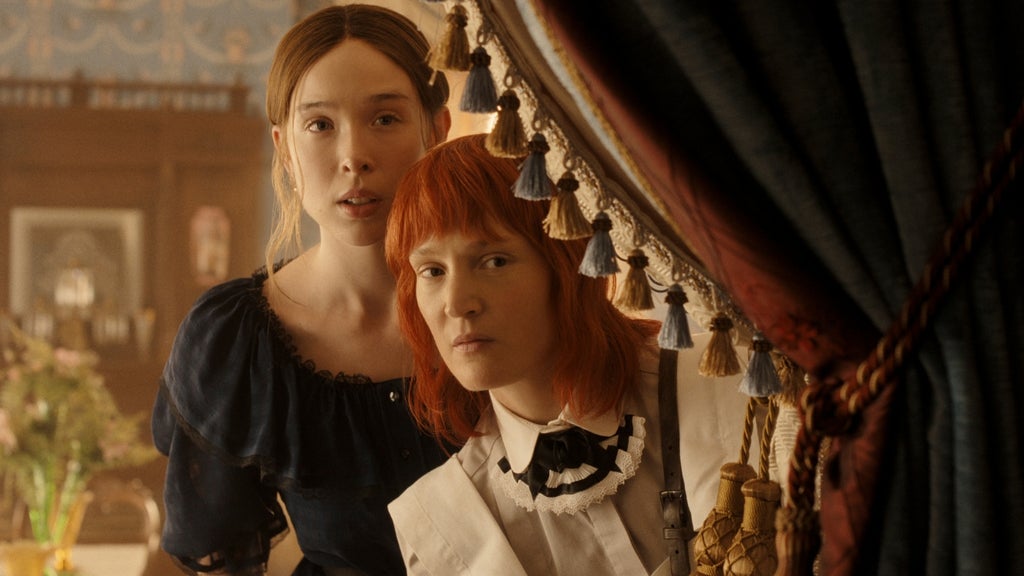 Two women peek out from behind a tasseled curtain: one in a maid's uniform with bright red hair and a bow collar, the other with dark hair in a navy ruffled dress.