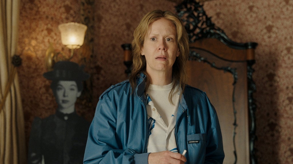 Woman with long blonde-brown hair and a startled expression stands in a blue jacket in an ornate wallpapered room with a black-and-white cutout figure behind her.