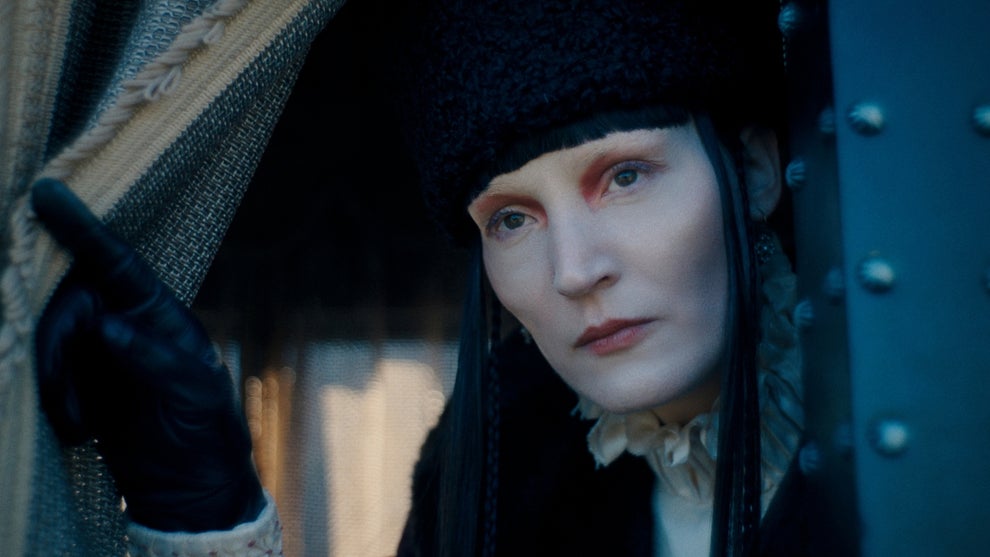 A pale-faced woman with dark fur hat, black gloves, and dramatic red-rimmed eye makeup peers out from behind a curtain.