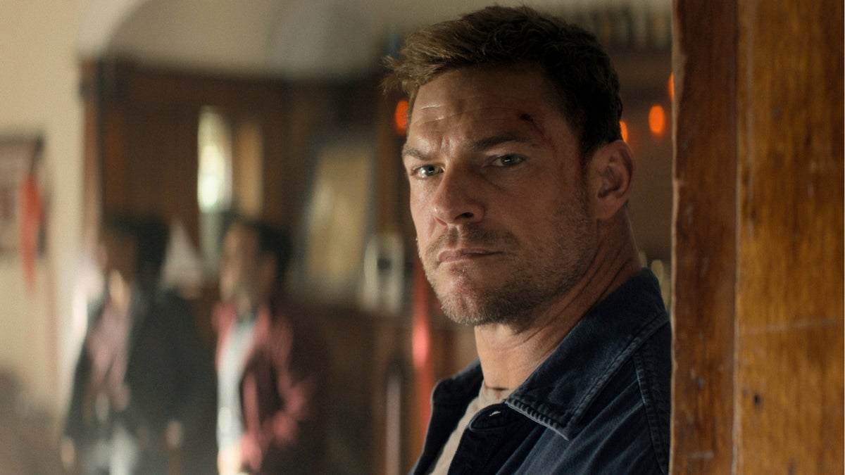 Alan Ritchson as Jack Reacher leans against a wooden doorframe, looking ahead past the camera