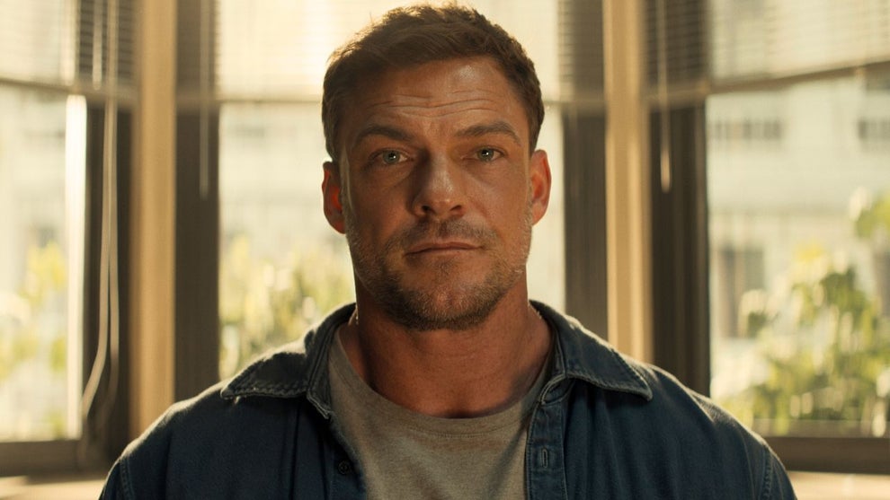 Alan Ritchson, in character as Jack Reacher, stares directly at the camera with a serious expression. He has short brown hair and light stubble, and wears a gray T-shirt under an open denim shirt. He stands in front of tall windows with blinds, softly backlit by daylight.