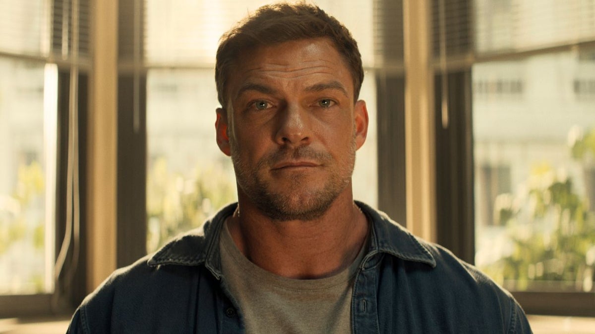 Alan Ritchson, in character as Jack Reacher, stares directly at the camera with a serious expression. He has short brown hair and light stubble, and wears a gray T-shirt under an open denim shirt. He stands in front of tall windows with blinds, softly backlit by daylight.