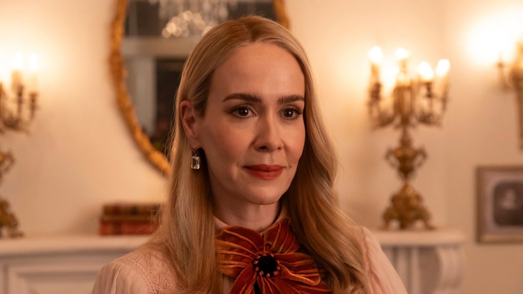 Blonde woman with wavy hair and a rust-colored bow blouse stands in a warmly lit room with gold candelabras and an ornate mirror behind her.