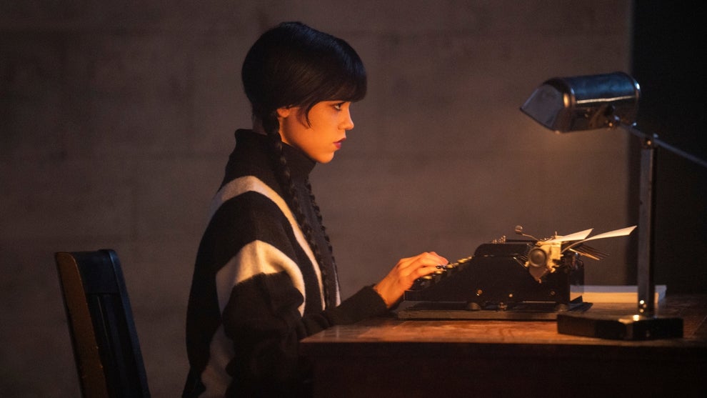 Jenna Ortega as Wednesday Addams sits in profile at a wooden desk using her typewriter. She has dark hair with bangs and wears a black and white sweater.