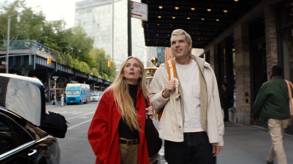 "A man and woman walk side by side on a busy city sidewalk near a building entrance, both looking upward; trees, cars, and people are visible in the urban background."
