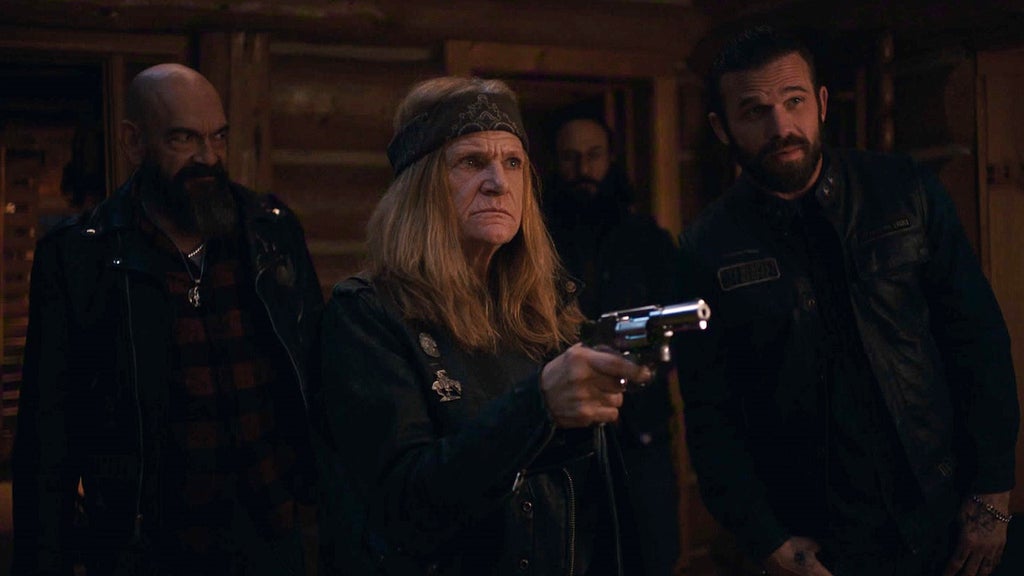 "Four people in dark biker attire stand inside a dimly lit wooden cabin, one older woman in the foreground holds a silver revolver, others look on tensely, suggesting a dramatic or confrontational moment."