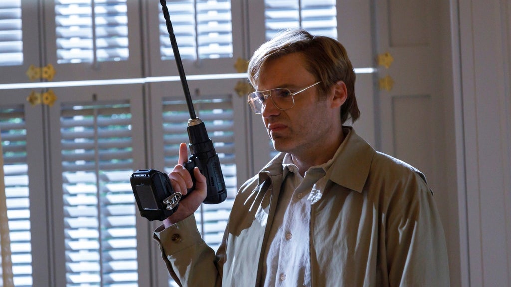 Seth Gabel as serial killer Jeffrey Dahmer in AHS 13, standing in front of blinds while holding a drill