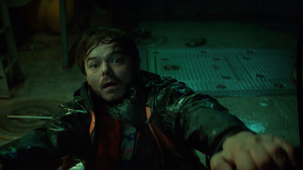 "A frightened man in a raincoat clings to a metal railing inside a dimly lit, industrial room with wet floors and scattered tools, suggesting tension or danger in the environment."