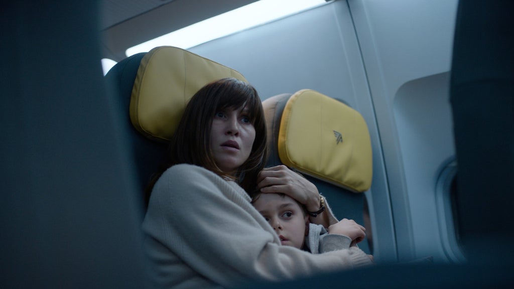 On a dimly lit airplane, a frightened woman with dark bangs and a cream sweater sits in a seat with a yellow headrest, holding a young boy protectively against her chest as they both stare ahead.