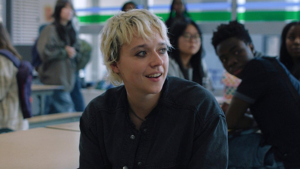A teenager with short bleached hair sits in a school room with a faint smile as she looks to the right. Students can be seen in the background sitting at tables.