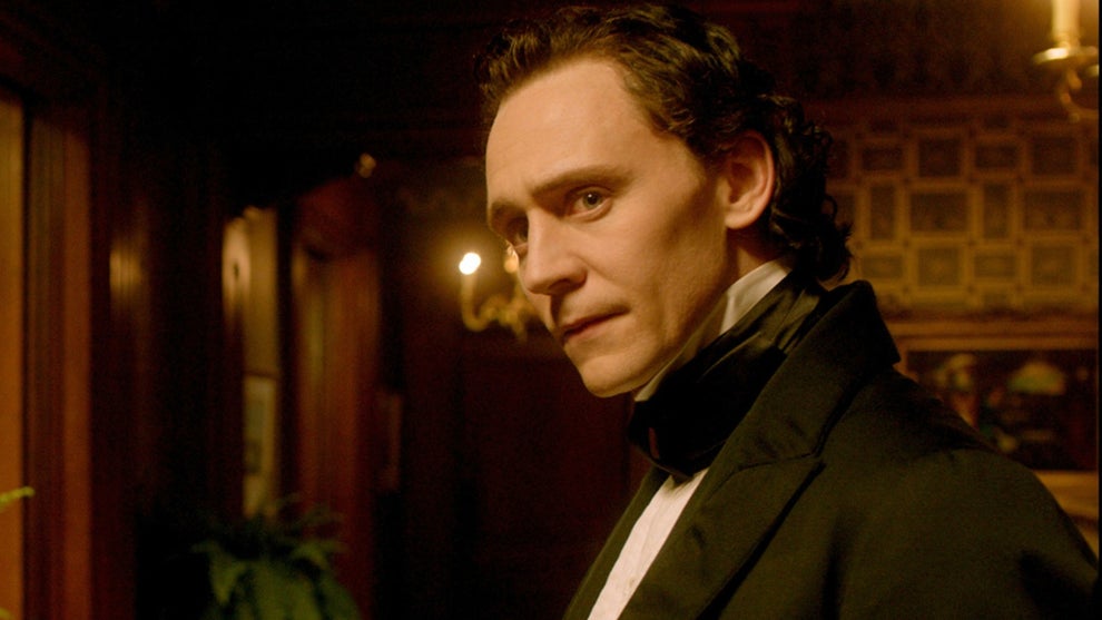 Tom Hiddleston wears 19th century attire in a black jacket and white collared shirt with his hair slicked back. An ornate, wood-paneled room can be seen behind him.