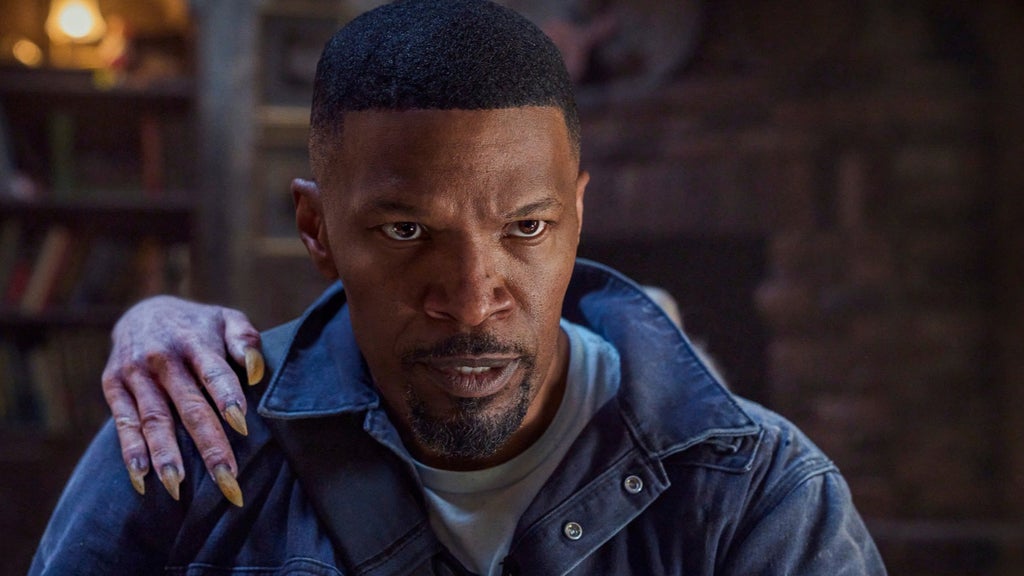 Jamie Foxx with a close-cropped haircut and goatee, wearing a denim jacket over a white T-shirt, stares warily off-camera. A gnarled, gray hand with long, yellowed claws rests on his shoulder from behind. The background is a dim, warmly lit room with bookshelves.