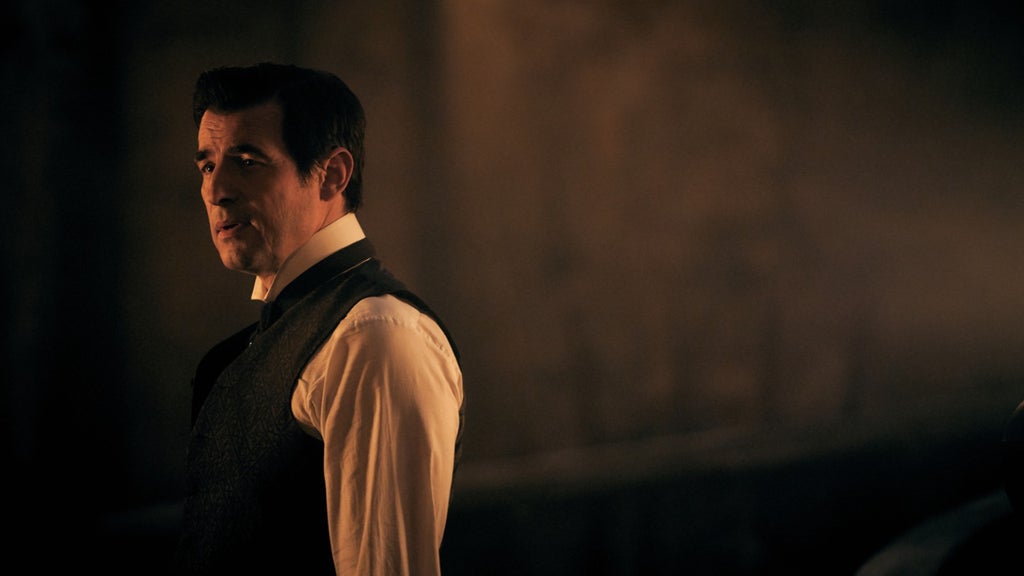 A dark-haired man in a white dress shirt, dark tie and patterned black waistcoat stands in a dim, amber-lit room, glancing to the left with a wry, knowing expression as if speaking. Warm light catches one side of his face against a hazy, shadowy background.