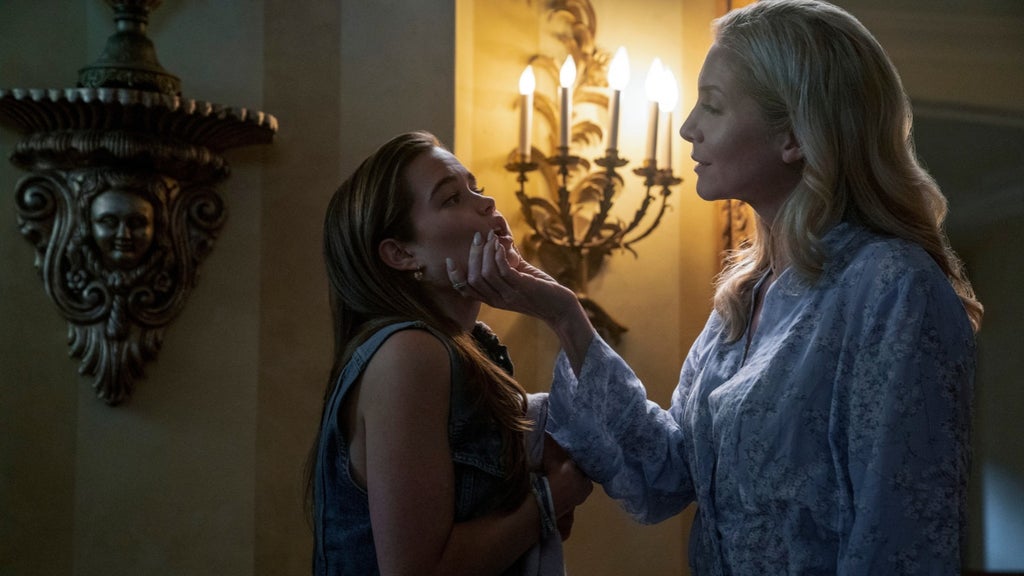 In a dim, ornately decorated hallway, a blonde woman in a floral blue blouse cups the chin of a young woman in a sleeveless denim top, who looks up at her uneasily. A lit candle-style wall sconce glows between them, beside a carved wall shelf with a face sculpted into it.
