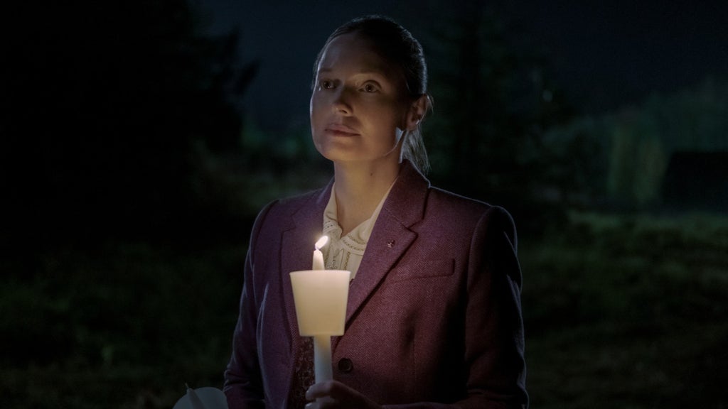 Samantha Sloyan in Midnight Mass, seen with her hair pulled back, stands outdoors at night, holding a lit candle in a paper drip guard as if at a vigil. She wears a burgundy herringbone blazer over a cream blouse with a lace collar, and gazes ahead with a solemn, expression. The candlelight softly illuminates her face against a dark background.