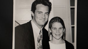"A black-and-white photo showing a man in a suit and tie smiling next to a young girl with braces. They are indoors, in front of a white wall with window panes. The atmosphere appears warm and friendly."